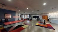This single-floor amenity space is thoughtfully designed to serve as a dynamic hub for recreation, relaxation, and productivity. The layout encompasses several distinct zones, each offering a unique function within a cohesive modern aesthetic.

The centerpiece of the space is a spacious game room featuring polished concrete floors, a full-size billiards table with a sleek contemporary frame, and a foosball table. Colorful geometric area rugs add vibrant accents throughout, complementing the room's bold blue accent wall adorned with framed artwork. A wall-mounted flat-screen TV and a pair of red accent chairs create a comfortable media viewing area nearby.

Adjacent to the game room is a lounge area furnished with a large dark leather sectional sofa, a glass coffee table, and views through expansive floor-to-ceiling windows overlooking an outdoor courtyard. A chalkboard wall provides a community messaging feature within this zone.

The fully equipped kitchen includes stainless steel appliances — notably a large built-in refrigerator and oven — ample cabinetry, a central island with a stone countertop, a wall-mounted pot rack, and pendant lighting, making it well-suited for group gatherings and events.

A dedicated business center offers computer workstations along a built-in desk, glass-top tables with red and chrome seating, a printer, and framed artwork, providing a functional workspace within the amenity suite.