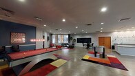 This single-floor amenity space is thoughtfully designed to serve as a dynamic hub for recreation, relaxation, and productivity. The layout encompasses several distinct zones, each offering a unique function within a cohesive modern aesthetic.

The centerpiece of the space is a spacious game room featuring polished concrete floors, a full-size billiards table with a sleek contemporary frame, and a foosball table. Colorful geometric area rugs add vibrant accents throughout, complementing the room's bold blue accent wall adorned with framed artwork. A wall-mounted flat-screen TV and a pair of red accent chairs create a comfortable media viewing area nearby.

Adjacent to the game room is a lounge area furnished with a large dark leather sectional sofa, a glass coffee table, and views through expansive floor-to-ceiling windows overlooking an outdoor courtyard. A chalkboard wall provides a community messaging feature within this zone.

The fully equipped kitchen includes stainless steel appliances — notably a large built-in refrigerator and oven — ample cabinetry, a central island with a stone countertop, a wall-mounted pot rack, and pendant lighting, making it well-suited for group gatherings and events.

A dedicated business center offers computer workstations along a built-in desk, glass-top tables with red and chrome seating, a printer, and framed artwork, providing a functional workspace within the amenity suite.