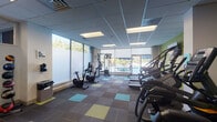 Located at 110 W 39th St in Baltimore, MD, this single-floor fitness center is thoughtfully designed to support a well-rounded workout experience. The space is anchored by a vibrant color palette, with bold green accent walls displaying motivational quotes and wall art, complemented by a multi-toned carpet tile floor in shades of gray, teal, and yellow-green that adds energy to the environment. A suspended ceiling with recessed panel lighting ensures the space is consistently well-lit throughout.

The cardio zone is well-equipped with treadmills, stair climbers, ellipticals, and stationary cycling bikes — several featuring individual display screens for an engaging workout. A wall-mounted flat-screen television is positioned above the treadmill area for added entertainment. Strength and flexibility training is also supported, with a cable machine, free weights, and stability balls neatly organized along the walls.

Large floor-to-ceiling windows and a glass sliding door line one side of the room, flooding the space with natural light and offering direct views of an outdoor pool and patio area. Frosted privacy film on a portion of the windows balances openness with privacy. The overall ambiance is clean, modern, and motivating — a functional and welcoming facility for daily fitness routines.