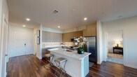 This contemporary single-floor residence showcases a thoughtfully designed open-concept layout with high-end finishes throughout. The kitchen is a standout feature, offering two-tone cabinetry in warm wood and cool grey tones, a large quartz waterfall island with bar seating, stainless steel appliances including a gas range and French door refrigerator, and a classic tile backsplash. Rich, dark hardwood flooring flows seamlessly through the main living areas, adding warmth and elegance.

The living room is bright and inviting, benefiting from natural light through large sliding glass doors that open to a private balcony with neighborhood views. A ceiling fan adds to the comfort of the space. Adjacent to the kitchen, a dedicated laundry area provides added convenience.

The primary bedroom is generously sized, featuring plush carpeting, a ceiling fan, a large window overlooking the outdoors, and direct access to a well-appointed en-suite bathroom. The bathroom is finished with marble-look tile walls in the glass-enclosed walk-in shower, a modern floating vanity with an undermount sink, and clean tile flooring. A spacious walk-in closet with multi-level shelving and hanging rods completes the primary suite, offering ample and well-organized storage.
