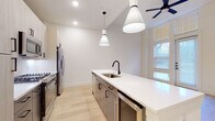 This single-floor residence showcases a clean, contemporary design with an open-concept layout that maximizes both function and flow. The kitchen serves as the centerpiece of the space, anchored by a generously sized white quartz island equipped with a stainless steel sink and matte black faucet. Elegant pendant lights hang above, adding a refined touch. Light-toned flat-panel cabinetry extends to the ceiling, providing ample storage, while a full suite of stainless steel appliances — including a French door refrigerator with ice dispenser, gas range, and built-in microwave — completes the well-equipped culinary space. A white subway tile backsplash and under-cabinet lighting add subtle detail. Light hardwood flooring runs throughout the main living areas, lending warmth and continuity. The living area benefits from tall, floor-to-ceiling windows and a glass door that allows natural light to pour in, creating an airy and open atmosphere. A ceiling fan adds comfort to this bright space. The bathroom is finished with large-format light tile flooring and features both a glass-enclosed shower and a separate soaking tub, offering a clean and functional layout. Throughout the residence, a neutral white and warm wood palette creates a modern, cohesive aesthetic.