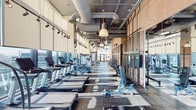 Located at 484 Spadina Avenue in Toronto's vibrant Kensington-Chinatown neighbourhood, this fitness facility offers a comprehensive, single-floor gym experience defined by its industrial-chic aesthetic and abundant natural light. Floor-to-ceiling steel-framed windows line the perimeter, flooding the space with sunlight and providing sweeping views of the Toronto skyline — a striking backdrop for any workout.

The main cardio floor is an elongated, open-plan area featuring a long row of treadmills and other cardio machines arranged along the window wall. Exposed ductwork and pendant lighting overhead contribute to the space's urban industrial character, while dark rubber flooring and track lighting add functionality and style.

A dedicated strength training zone houses power racks, weight plates, and resistance equipment, offering ample room for free-weight and functional training. Glass partitions divide sections of the gym, maintaining an open and airy feel while defining distinct workout areas.

A separate cycling studio features spin bikes on hardwood flooring, mirrored walls, and large steel-framed glass walls that connect visually to the surrounding fitness floor. A dedicated mind and body room provides a calm, wood-panelled retreat equipped with exercise mats and a wall-mounted display for guided workouts, ideal for yoga, stretching, and recovery sessions.