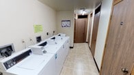 This shared laundry facility is a practical, well-organized space designed to serve the needs of its users efficiently. The room features a row of top-load washing machines and matching dryers lined along one wall, providing ample capacity for multiple loads of laundry. The machines are arranged in a clean, accessible layout, making the laundry process straightforward and convenient.

The flooring consists of neutral-toned tile, offering durability and ease of maintenance. The walls are painted in a light, neutral color, contributing to the bright and functional atmosphere of the room. Posted on the walls are a clearly visible set of laundry room rules and an operating instructions guide, ensuring users have all the information they need at a glance.

At the far end of the room, two wooden doors with transom windows above lead to additional areas, adding depth to the space. Overhead fluorescent lighting ensures the room is well-lit throughout. The overall ambiance is clean, utilitarian, and purpose-driven, reflecting a space that prioritizes functionality and ease of use for all who utilize it.
