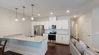This two-story home offers a thoughtfully designed layout with contemporary finishes and a cohesive aesthetic across both levels. The main living area centers around an open-concept arrangement where the kitchen, dining, and living spaces flow seamlessly together. Rich dark hardwood flooring anchors the space throughout the main level, contrasting beautifully with the light, neutral wall tones.

The kitchen is a standout feature, outfitted with crisp white shaker cabinetry, granite countertops, a subway tile backsplash, and a full suite of stainless steel appliances. A large central island with an undermount sink and pendant lighting above provides both additional prep space and casual seating. The adjacent dining area benefits from natural light through a window overlooking the exterior, while the living room offers a comfortable, relaxed setting with a ceiling fan and additional window views.

The bedrooms are carpeted for comfort and each include ceiling fans and windows with outdoor views. Bathrooms are finished with white cabinetry, granite countertops, and wide vanity mirrors. A dedicated laundry room includes wire shelving, washer/dryer hookups, and a gas connection. The overall design balances modern style with functional, everyday livability.