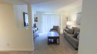 This single-floor space showcases a welcoming living area designed with a neutral, understated aesthetic. The room is finished with wall-to-wall plush carpeting in a warm beige tone, contributing to a comfortable and inviting atmosphere. Soft, cream-colored walls complement the light palette throughout the space.

A prominent sliding glass door dressed with vertical blinds serves as a key architectural feature, allowing an abundance of natural light to fill the room while also providing potential access to an outdoor area. A floor lamp and table lamp offer additional ambient lighting options for the evenings.

The living area is thoughtfully laid out to accommodate a sofa with decorative accent pillows, a matching armchair, and a dark wood coffee table with a lower storage shelf—providing both style and function. A low-profile dresser or media console with multiple drawers is positioned along one wall, topped with decorative accessories, and a framed mirror or artwork is mounted nearby. Structural columns add architectural character to the space. Overall, the space presents a clean, functional layout with a calm, neutral ambiance suitable for everyday living.