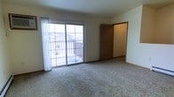 This single-story space features a generously sized main room finished with neutral wall-to-wall carpeting that lends a comfortable, understated character to the interior. The room is anchored by a large sliding glass door fitted with sheer white curtains, which allows abundant natural light to fill the space throughout the day. The sliding door provides direct access to an outdoor balcony enclosed by a classic railing, overlooking a residential neighborhood — a notable feature that brings an open, airy quality to the room. A medium-toned wooden door with a traditional six-panel design leads to an adjacent area, likely a hallway or additional room, adding functional connectivity to the layout. The walls are finished in a clean, neutral tone that complements the light carpeting and contributes to the overall bright and open ambiance. Baseboards run along the perimeter, providing a clean transition between the walls and flooring. The space presents a straightforward, practical layout well-suited to a variety of uses, with its combination of natural light, outdoor access, and neutral finishes creating a welcoming and versatile environment.