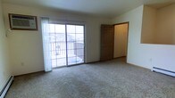 This single-story space features a generously sized main room finished with neutral wall-to-wall carpeting that lends a comfortable, understated character to the interior. The room is anchored by a large sliding glass door fitted with sheer white curtains, which allows abundant natural light to fill the space throughout the day. The sliding door provides direct access to an outdoor balcony enclosed by a classic railing, overlooking a residential neighborhood — a notable feature that brings an open, airy quality to the room. A medium-toned wooden door with a traditional six-panel design leads to an adjacent area, likely a hallway or additional room, adding functional connectivity to the layout. The walls are finished in a clean, neutral tone that complements the light carpeting and contributes to the overall bright and open ambiance. Baseboards run along the perimeter, providing a clean transition between the walls and flooring. The space presents a straightforward, practical layout well-suited to a variety of uses, with its combination of natural light, outdoor access, and neutral finishes creating a welcoming and versatile environment.
