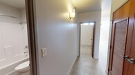This single-floor unit offers a well-organized layout comprising a hallway, a walk-in closet, and a full bathroom. The open living area is a standout feature, highlighted by large, floor-to-ceiling windows framed in bold red, flooding the space with natural light and offering expansive views of the surrounding cityscape and tree-covered hillside. The concrete floors throughout lend an urban, industrial aesthetic to the space.

The kitchen is efficiently designed along a single wall, featuring white cabinetry that extends to the ceiling for ample storage, a yellow ceramic tile backsplash that adds a vibrant pop of color, stone countertops, a double sink, and a full suite of white appliances including a refrigerator, electric range, dishwasher, and built-in microwave.

The walk-in closet is generously sized and fully equipped with multi-tiered wire shelving and hanging systems on three walls, providing extensive storage and organization options. The bathroom includes a full soaking tub with an overhead shower, white surround walls, yellow tile accents that echo the kitchen's color palette, and a chrome towel rack. The hallway connects all areas efficiently and is illuminated by a wall-mounted light fixture, completing this compact yet functional space.