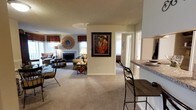 This single-floor residence showcases an open-concept design that connects the living, dining, and kitchen areas in a cohesive, functional layout. The spacious living room is anchored by a classic fireplace framed in white, providing a warm focal point. Neutral beige walls and soft wall-to-wall carpeting throughout the main areas create a calm, inviting atmosphere. Natural light flows in generously through multiple windows, each dressed with decorative valances in warm earth tones that add a traditional character to the space. A sunburst mirror and framed artwork lend decorative personality to the walls.

Adjacent to the living area, a breakfast bar with granite-style countertops and barstool seating offers a convenient casual dining option, seamlessly connecting to what appears to be a well-appointed kitchen. The textured ceiling adds depth overhead, while the open sightlines between rooms enhance the overall sense of spaciousness. A hallway visible in the background suggests additional private rooms beyond, contributing to a well-organized and comfortable floor plan suited for everyday living.