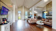 This single-story space offers a generous, open-plan layout designed to accommodate a variety of functions, from professional workstations to comfortable lounge and dining areas. The architectural design is immediately striking, with dramatically vaulted ceilings that rise to impressive heights throughout the main area, supported by white structural columns that define zones without enclosing them. Rich, dark hardwood flooring runs throughout, providing a warm and cohesive aesthetic.

The main area serves multiple purposes seamlessly. A welcoming lounge features leather sofas and a round coffee table, positioned near large windows that flood the space with natural light and offer views of the surrounding greenery. Adjacent to the lounge, a dining area centers around a solid wood farm table beneath a statement pendant light. A floor-to-ceiling stacked stone fireplace anchors this section, adding a striking focal point and a sense of warmth.

A dedicated workspace includes desks and workstations, while a mounted flat-screen TV and a coffee station along one wall add to the space's functional and communal character. The kitchen is fully equipped with warm wood cabinetry, a breakfast bar peninsula, black appliances, and a tall window that brings in additional natural light, completing this versatile and well-appointed space.