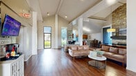 This single-story space offers a generous, open-plan layout designed to accommodate a variety of functions, from professional workstations to comfortable lounge and dining areas. The architectural design is immediately striking, with dramatically vaulted ceilings that rise to impressive heights throughout the main area, supported by white structural columns that define zones without enclosing them. Rich, dark hardwood flooring runs throughout, providing a warm and cohesive aesthetic.

The main area serves multiple purposes seamlessly. A welcoming lounge features leather sofas and a round coffee table, positioned near large windows that flood the space with natural light and offer views of the surrounding greenery. Adjacent to the lounge, a dining area centers around a solid wood farm table beneath a statement pendant light. A floor-to-ceiling stacked stone fireplace anchors this section, adding a striking focal point and a sense of warmth.

A dedicated workspace includes desks and workstations, while a mounted flat-screen TV and a coffee station along one wall add to the space's functional and communal character. The kitchen is fully equipped with warm wood cabinetry, a breakfast bar peninsula, black appliances, and a tall window that brings in additional natural light, completing this versatile and well-appointed space.