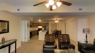 This single-floor residence offers an open-concept layout that effortlessly connects the main living areas, creating a cohesive and functional living environment. The living room is comfortably furnished with leather seating and accent chairs, anchored by a ceiling fan that adds both style and airflow to the space. Neutral-toned carpeting runs throughout, lending warmth and continuity across the open floor plan. The kitchen is well-appointed with dark cabinetry, black appliances including a refrigerator and range, and a white central island that serves as both a prep space and a casual gathering point. Overhead lighting and a secondary ceiling fan keep the kitchen and adjoining areas bright and comfortable. Adjacent to the kitchen, a dedicated dining area accommodates a full dining table and chairs, making it ideal for everyday meals or entertaining guests. The layout benefits from a smooth flow between zones, with doorways leading to additional rooms beyond. The overall ambiance is practical and inviting, with a cohesive design aesthetic defined by dark accents, neutral tones, and an efficiently organized use of space throughout the single level.