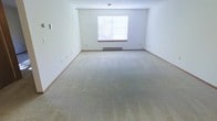 This single-floor space presents a generously sized open room with a clean, minimalist aesthetic. The interior is finished with wall-to-wall neutral-toned carpeting that extends throughout, providing a soft and understated foundation underfoot. The walls are painted in a crisp white, contributing to a bright and airy atmosphere that makes the room feel open and versatile.

A centrally positioned window fitted with horizontal blinds allows natural light to filter into the space, illuminating the room while offering adjustable privacy. Beneath the window sits a baseboard heater, indicating a dedicated heating system integrated directly into the room's perimeter. The white painted baseboard trim runs along the base of the walls, providing a clean boundary between the walls and carpeted floor.

Multiple electrical outlets are distributed along the walls, offering practical placement for various uses. The room's generous proportions and open layout make it highly adaptable, suitable for a range of furnishing configurations. The overall ambiance is neutral, clean, and move-in ready, offering a blank canvas for personalization.