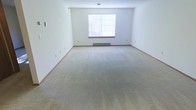 This single-floor space presents a generously sized open room with a clean, minimalist aesthetic. The interior is finished with wall-to-wall neutral-toned carpeting that extends throughout, providing a soft and understated foundation underfoot. The walls are painted in a crisp white, contributing to a bright and airy atmosphere that makes the room feel open and versatile.

A centrally positioned window fitted with horizontal blinds allows natural light to filter into the space, illuminating the room while offering adjustable privacy. Beneath the window sits a baseboard heater, indicating a dedicated heating system integrated directly into the room's perimeter. The white painted baseboard trim runs along the base of the walls, providing a clean boundary between the walls and carpeted floor.

Multiple electrical outlets are distributed along the walls, offering practical placement for various uses. The room's generous proportions and open layout make it highly adaptable, suitable for a range of furnishing configurations. The overall ambiance is neutral, clean, and move-in ready, offering a blank canvas for personalization.