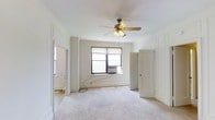 This single-floor space presents a clean, understated aesthetic defined by classic architectural details and a neutral color palette throughout. The main living area is finished with plush wall-to-wall carpeting in a soft, light tone, creating a warm and comfortable atmosphere underfoot. Crisp white walls are accented by traditional paneled molding and wide door casings, lending the space a timeless character. A multi-blade ceiling fan with a built-in light fixture anchors the center of the room, providing both air circulation and ambient lighting. Natural light filters in through a set of windows fitted with roller shades and a wall-mounted air conditioning unit, ensuring year-round comfort. Multiple doorways connect the main room to adjacent spaces, suggesting a practical and well-considered layout that allows for easy flow between areas. The overall ambiance is bright, open, and move-in ready, with a straightforward design that provides a versatile backdrop suited to a variety of furnishing styles and uses. The space's architectural detailing and neutral finishes combine to create an environment that feels both welcoming and functional.