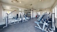 Located at 1469 Zepol Rd in Santa Fe, New Mexico, this fitness center offers a well-organized, single-room workout facility designed to accommodate a comprehensive range of exercise needs. The space features durable rubber flooring throughout, providing a safe and functional surface for both cardio and strength training activities.

The room is equipped with multiple cardio machines, including treadmills, ellipticals, and a stair climber, positioned along the windowed walls to take advantage of natural light streaming in from multiple directions. A wall-mounted air conditioning unit ensures comfortable year-round use.

The strength training area features a centrally located power rack with cable attachments and an integrated weight storage system, complemented by a selection of selectorized machines and adjustable benches. A dedicated dumbbell rack provides a range of free weights for versatile training options. Full-length mirrors along one wall enhance the sense of space and allow users to monitor their form during workouts.

Overhead, track lighting and ceiling fans provide ample illumination and air circulation. The white walls and light-toned ceiling contribute to a clean, airy atmosphere. Two access doors and multiple windows connect the space to its surroundings, ensuring the facility feels open and accessible despite its compact footprint.