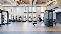 Located at 1301 Lebanon Pike in Nashville, TN, this contemporary fitness center offers a well-appointed, single-floor layout designed to accommodate a full range of workout experiences. The main exercise floor is expansive and open, featuring soaring double-height ceilings adorned with coffered details and large circular pendant light fixtures. Rows of floor-to-ceiling windows flood the space with natural light while offering views of the surrounding community. The flooring throughout the main area is a light-toned hardwood-style surface, contributing to the clean, modern aesthetic. A dedicated strength-training zone is delineated by elegant arched openings and equipped with power racks, a full dumbbell rack, adjustable benches, and cable machines. Cardio equipment, including treadmills, ellipticals, and stair climbers, is arranged along the windowed walls to maximize views and ventilation. Ceiling fans are integrated into the coffered ceiling design for added comfort. A separate studio space features warm natural wood paneling on the walls and ceiling, a wall-mounted display screen for guided workouts, and writable boards — ideal for group fitness classes or personal training sessions. The facility also includes a garage-door entry point that opens directly to the outdoors, seamlessly connecting the interior to the surrounding grounds.