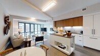 This thoughtfully designed single-floor residence showcases a contemporary open-concept layout that seamlessly connects the kitchen, dining, and living areas into one cohesive space. The kitchen is a standout feature, equipped with stainless steel Bosch appliances, a gas cooktop, and a combination of warm walnut upper cabinetry and crisp white lower cabinets. A central island with a dark countertop provides additional prep space and casual seating, all illuminated by under-cabinet lighting and a white subway tile backsplash. The open living area is bathed in natural light through expansive floor-to-ceiling windows, offering urban views and a bright, airy ambiance. The living room features neutral tones and contemporary furnishings, with a sliding barn door connecting to the adjacent spaces. Two well-proportioned bedrooms maintain the soft, neutral palette with warm wood accents and stylish framed artwork. A dedicated home office provides a quiet workspace complete with built-in desk and open shelving. The full bathroom impresses with floor-to-ceiling herringbone tile, a large frameless glass walk-in shower, and warm wood vanity cabinetry, delivering a polished, spa-like finish throughout.