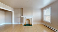 This single-floor space combines classic architectural details with functional design throughout. The living area serves as a standout feature, showcasing a decorative fireplace framed by a white wood mantel and an ornate surround of gold and green tiles set against a dark green tiled hearth — a distinctive focal point that adds character to the room. Light-toned hardwood flooring runs consistently throughout the space, lending warmth and cohesion to each room.

Multiple bedrooms offer comfortable proportions, each finished with the same hardwood flooring, neutral warm-toned walls, white baseboard trim, and baseboard heating units. Large windows with blinds allow natural light to fill the rooms while providing views of the surrounding neighborhood.

The kitchen is well-equipped with ample white cabinetry extending to the ceiling, dark countertops, a dark tile backsplash, a stainless steel sink, and hexagonal marble-style tile flooring — creating a clean, classic aesthetic.

The bathroom continues the classic theme with hexagonal marble-style floor tiles, a wall-mounted sink, a mirror, a bathtub with tile surround, and a window that brings in natural light, maintaining a bright and airy atmosphere.