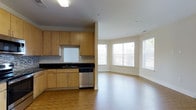 This single-floor space is designed with an open and functional layout that seamlessly connects the kitchen and living areas. The kitchen is outfitted with light maple cabinetry offering ample storage, dark granite countertops, a stainless steel dishwasher, and a built-in sink — providing a clean, modern aesthetic. The flooring throughout features warm hardwood, lending continuity and a polished look across the space.

Adjacent to the kitchen, the living area benefits from a curved bay window arrangement that spans a generous portion of the wall, inviting abundant natural light and creating an airy, welcoming atmosphere. The curved architectural detail adds a distinctive character to the overall layout.

A hallway connects additional areas of the unit, lined with crisp white walls, wide baseboards, and a paneled interior door with brushed nickel hardware. A wall-mounted HVAC vent is integrated neatly into the corridor. The neutral color palette throughout — creamy whites and warm wood tones — contributes to a bright, clean, and move-in-ready environment suited for comfortable everyday living.