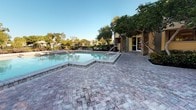 This outdoor pool area offers a well-appointed resort-style retreat designed for relaxation and recreation. The centerpiece is a generously sized swimming pool with clear blue water, framed by a broad, textured paver deck that provides ample space for sunbathing and socializing. The herringbone-pattern brick pavers extend throughout the entire deck, lending a warm, classic aesthetic underfoot.

Multiple chaise lounge chairs are positioned across the deck, offering comfortable spots to unwind in the sun or shade. The surrounding landscape is rich with mature trees, tropical palms, and manicured shrubbery, creating a lush, secluded atmosphere that enhances the sense of privacy. Additional outdoor seating areas with tables and chairs are tucked among the greenery, ideal for casual gatherings.

The adjacent building features a combination of green siding and stone veneer accents, with large glass doors providing direct access to the pool area. Exterior wall-mounted lighting adds ambiance for evening use. The pool includes built-in entry steps and is bordered by raised pool coping, giving it a clean, finished look. Under open skies and framed by mature canopy trees, this outdoor space delivers an inviting, park-like environment suited for leisurely days outdoors.