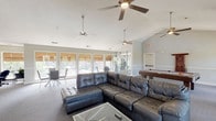 This single-floor community clubhouse is thoughtfully designed to serve a variety of social and recreational needs. The main gathering area is expansive and open, anchored by a large tufted leather sectional sofa and a glass-topped coffee table, creating a comfortable lounge atmosphere. Multiple ceiling fans are distributed throughout the space, complementing the vaulted ceiling that adds a sense of airiness and volume overhead. The flooring throughout is wall-to-wall carpet in a neutral gray tone, contributing to a clean and cohesive aesthetic.

Natural light flows generously through a broad bank of windows fitted with warm bamboo Roman shades, offering pleasant outdoor views while maintaining privacy. Doors opening to an outdoor balcony area extend the sense of openness further.

A pool table positioned toward one end of the main room serves as a focal point for recreation, while a designated fitness area with exercise equipment is accessible from the main space. A bar-height counter with sleek adjustable stools provides a casual seating option, and a dedicated workstation corner offers a quiet area for individual tasks.

The kitchen is well-appointed with warm oak cabinetry, dark granite-style countertops in a U-shaped layout, and a double sink — ideal for catering and community events. Additional windows in the kitchen bring in natural light. A dining area with a glass-top table and modern chairs rounds out this multifunctional clubhouse space.