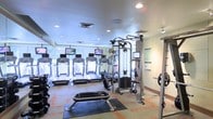 This fitness center is a thoughtfully designed single-floor facility offering a comprehensive range of exercise equipment to accommodate various workout needs. A row of treadmills lines the perimeter near floor-to-ceiling windows, each unit equipped with its own individual monitor for entertainment during cardio sessions. The large windows allow abundant natural light to fill the space while providing pleasant views of outdoor greenery, creating an inviting and energizing atmosphere.

At the center of the room, a multi-station cable machine serves as the focal point for strength and resistance training, complemented by free weights and a flat utility bench positioned on protective rubber matting. The layout is well-organized, providing ample circulation space between equipment for safe and comfortable use.

The facility features a light-toned ceiling with recessed lighting, ensuring consistent illumination throughout. Neutral-colored walls with informational postings keep the environment clean and functional. The patterned flooring adds a subtle design element while providing durability underfoot. Overall, the space strikes a balance between practicality and comfort, making it a fully functional fitness destination.