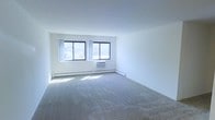 This single-floor space features a clean, minimalist design with neutral white walls and wall-to-wall carpeting in a light gray tone, creating a simple and functional interior. The main living area is generously sized, offering an open layout that provides flexibility for various furniture arrangements. Two large windows along the exterior wall allow ample natural light to fill the room, brightening the space throughout the day. The windows also offer views of the surrounding exterior area. Climate control is addressed through a combination of a wall-mounted air conditioning unit positioned beneath the windows and baseboard heating that runs along the perimeter of the room, ensuring year-round comfort. The ceiling is standard height with a smooth, white finish that complements the bright, airy feel of the room. The overall ambiance is straightforward and practical, with a move-in ready aesthetic suited for those who prefer a blank canvas. The space maintains a clean, uncluttered character that makes it easy to personalize according to individual preferences and needs.