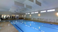 This single-floor indoor aquatic facility centers around a generously sized lap pool, designed to accommodate serious swimmers and recreational users alike. The pool is clearly divided into multiple lanes using floating lane dividers, ensuring an organized swimming environment. The water appears clean and well-maintained, reflecting the warm ambient lighting throughout the space.

The facility features notably high ceilings with distinctive rectangular acoustic panels suspended overhead, contributing to both sound management and visual interest. A row of clerestory windows lines the upper portion of the walls, allowing natural light to filter in while maintaining privacy at the pool level. Wall-mounted sconces provide additional warm, even illumination throughout the space.

The surrounding pool deck is finished in light-toned tile, offering a clean and slip-resistant surface. The walls are accented with stone-like tile wainscoting, adding a refined, upscale touch to the overall aesthetic. Safety equipment, including a life ring, is visibly mounted on the wall. A glass-door entrance at one end provides access to adjacent facilities. The overall ambiance is calm, clean, and professionally maintained.
