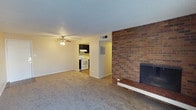 This single-floor space is centered around a spacious living area finished with neutral wall-to-wall carpeting that lends a warm, understated tone throughout. The most striking architectural feature is the full-height brick fireplace wall, constructed in rich red brick with a raised tile hearth, serving as a bold focal point that anchors the room. A ceiling fan with an integrated light fixture is centrally positioned overhead, providing both air circulation and ambient lighting to the open living space. The textured ceiling adds a classic residential character consistent with the overall design.

An open passageway connects the living area directly to the kitchen, which features dark cabinetry, a white electric range with an overhead range hood, and light-colored flooring that contrasts with the carpeted living space. This layout creates a functional flow between the two areas. A utility panel is visible along the shared wall between the kitchen and living room, alongside a ceiling-mounted HVAC vent, indicating built-in climate control. The overall design is practical and straightforward, combining durable materials with classic residential details.