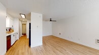 This single-floor residence offers a clean, modern aesthetic throughout its thoughtfully laid-out interior. The open living area is a standout feature, showcasing light-toned hardwood-style flooring, crisp white walls, and a textured ceiling. A ceiling fan with integrated lighting anchors the living space, while a large window with blinds floods the room with natural light and provides views of the surrounding neighborhood. A wall-mounted heater vent adds a practical touch to the open area.

The kitchen is positioned adjacent to the living space, partially separated by a structural half-wall that adds definition without closing off the room. It features white shaker-style cabinetry with matte black hardware, light countertops, a stainless steel sink with a black faucet, and black appliances including a refrigerator and dishwasher, all set atop the same warm hardwood-style flooring.

The bathroom is bright and contemporary, outfitted with a white vanity, stone-topped countertop, a large mirror, matte black fixtures, and a combined tub and shower with clean white surround. The bedroom offers plush carpeting, a window with greenery views, a textured ceiling, and a wall vent, creating a comfortable and quiet retreat. The hallway connects all rooms with consistent, light-filled finishes throughout.