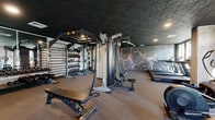 This modern fitness center is thoughtfully designed to accommodate a wide range of workout styles across several distinct zones. The space is unified by a bold aesthetic, featuring geometric-patterned ceilings finished in deep black, rubber flooring throughout, and dramatic athletic murals adorning the walls, creating an energizing atmosphere for exercise.

The strength training area is comprehensively outfitted with a multi-functional power rack, an extensive collection of kettlebells and free weights organized on wall-mounted storage racks, a cable machine, and adjustable weight benches. A dedicated zone provides additional space for bench pressing and barbell work, with neatly arranged weight plates and barbells close at hand.

The cardio section offers multiple treadmills with integrated touchscreen displays, elliptical trainers, a stair climber, stationary bikes including Echelon smart bikes, and a rowing machine. Large windows line this area, allowing natural light to fill the space. Full-length mirrors in one section enhance the sense of openness.

Additional amenities include a water fountain station, glass-panel entry doors, and ample open floor space for stretching or functional training. The overall design balances high-performance functionality with a sleek, contemporary aesthetic.