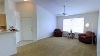 This single-floor space showcases a welcoming and open living area designed with both comfort and functionality in mind. The room's most striking architectural feature is its vaulted ceiling, which lends a sense of height and airiness to the interior. A ceiling fan is centrally mounted, providing efficient air circulation throughout the space.

The living area is finished with wall-to-wall carpeting in a warm neutral tone, creating a soft and inviting atmosphere underfoot. A large window positioned along one wall allows an abundance of natural light to pour in, brightening the entire room and offering a connection to the outdoors. The walls are painted in a clean, crisp white, further enhancing the bright and open feel of the space.

A solid entry door with a secure deadbolt provides direct access to the living area, offering both convenience and peace of mind. The open layout provides ample room for a variety of furniture arrangements, making it highly versatile for everyday living. Overall, the space combines practical design elements with an airy, light-filled ambiance that creates a comfortable and inviting environment.