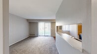 This single-story unit offers an efficient and thoughtfully arranged layout encompassing a living room, dining room, kitchen, and one bedroom. The living room is a welcoming, carpeted space characterized by neutral gray walls and a large sliding glass door fitted with vertical blinds, allowing abundant natural light and providing outdoor views. A half-wall partition separates the living area from the adjoining dining and kitchen spaces, creating an open yet defined floor plan.

The galley-style kitchen features warm-toned wood cabinetry lining both walls, light countertops, a double sink, and hardwood-style flooring. White appliances, including a range and refrigerator, are neatly integrated into the cabinetry. A pass-through window connects the kitchen visually to the dining area, enhancing the open feel.

The dining area transitions naturally between the kitchen and living spaces, maintaining the cohesive, open character of the unit. The bedroom offers a comfortable, carpeted retreat with a window dressed in vertical blinds that filters natural light, a closet, and neutral walls. Throughout the unit, a consistent palette of soft grays and whites contributes to a clean, calm ambiance.