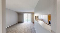 This single-story unit offers an efficient and thoughtfully arranged layout encompassing a living room, dining room, kitchen, and one bedroom. The living room is a welcoming, carpeted space characterized by neutral gray walls and a large sliding glass door fitted with vertical blinds, allowing abundant natural light and providing outdoor views. A half-wall partition separates the living area from the adjoining dining and kitchen spaces, creating an open yet defined floor plan.

The galley-style kitchen features warm-toned wood cabinetry lining both walls, light countertops, a double sink, and hardwood-style flooring. White appliances, including a range and refrigerator, are neatly integrated into the cabinetry. A pass-through window connects the kitchen visually to the dining area, enhancing the open feel.

The dining area transitions naturally between the kitchen and living spaces, maintaining the cohesive, open character of the unit. The bedroom offers a comfortable, carpeted retreat with a window dressed in vertical blinds that filters natural light, a closet, and neutral walls. Throughout the unit, a consistent palette of soft grays and whites contributes to a clean, calm ambiance.