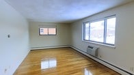 This single-floor space offers a clean and functional layout with a bright, open feel throughout. The main living area is highlighted by gleaming, polished hardwood floors that reflect natural light and lend the room a warm, inviting character. Two windows — one facing the front and one on the adjacent wall — allow ample daylight to fill the space while offering views of the surrounding neighborhood. The walls are finished in a neutral, light tone that complements the overall brightness of the interior. A wall-mounted HVAC unit provides convenient, independent climate control for year-round comfort. Baseboard heating runs along the perimeter of the room, adding a supplementary heating option. The ceiling features a smooth, white finish that contributes to the airy ambiance of the space. The room's open configuration provides flexibility for a variety of furniture arrangements and uses. Overall, the space presents a tidy, well-maintained interior with practical amenities and a straightforward design that emphasizes comfort and usability.
