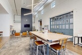 This generously sized single-floor commercial space is designed with a modern industrial aesthetic that balances open, collaborative areas with defined functional zones. The architecture is immediately distinguished by its dramatically high ceilings, which amplify the sense of volume throughout the main area and are accentuated by striking geometric linear pendant lights suspended overhead.

The primary gathering area features a mix of flooring — warm hardwood planks transitioning to patterned carpet — defining distinct seating zones. A large communal high-top table with bar-height seating serves as a central hub, surrounded by clusters of lounge chairs with side tables for more informal meetings. A signature blue-gray glazed garage-style door, branded "RUXTON," serves as a notable architectural focal point.

Large floor-to-ceiling windows flood the front lounge area with natural light and views of the surrounding streetscape. Exposed ductwork and brick-textured accent walls contribute to the space's industrial character, while white-painted walls keep the atmosphere bright and open.

A dedicated fitness room is equipped with treadmills, a stationary bike, a rowing machine, and a wall-mounted television, featuring its own hardwood flooring and exposed ventilation. Throughout, the design emphasizes functionality, openness, and a collaborative community atmosphere.