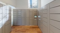 This package room is a well-organized, single-level space designed specifically for the secure receipt and retrieval of parcels. The room is lined with multiple banks of floor-to-ceiling gray smart parcel lockers, offering a wide range of compartment sizes to accommodate packages of varying dimensions. A centrally located digital kiosk interface is integrated into the locker system, enabling residents to receive notifications and retrieve their deliveries efficiently.

The room's interior features warm-toned hardwood flooring throughout, complementing the clean, white walls and crisp white baseboards. Recessed ceiling lights provide consistent, even illumination across the space. Natural light is introduced through glass-panel entry doors and high-positioned windows, adding brightness to the otherwise functional environment.

Access is provided through a solid paneled door alongside a glass-inset entry door, offering visibility into the space from the exterior. The layout is open and unobstructed, allowing for comfortable navigation around the locker banks. The overall design is clean and utilitarian, prioritizing functionality while maintaining a polished, well-maintained aesthetic consistent with a modern residential or mixed-use building amenity space.