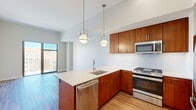 This well-appointed one-bedroom, one-bathroom residence offers a thoughtfully designed layout across a single floor. The open-concept kitchen and living area serves as the heart of the home, featuring warm-toned wood cabinetry, a striking quartz waterfall island with an undermount sink, and a full suite of stainless steel appliances including a gas range, microwave, and refrigerator. A white subway tile backsplash and globe pendant lights suspended from soaring ceilings add a polished, modern touch. Light hardwood-style flooring flows throughout the main living area and into the bedroom, creating a cohesive aesthetic. The living space opens directly to a private balcony through floor-to-ceiling glass sliding doors, filling the room with abundant natural light and offering open urban views. The bedroom is a comfortable retreat with large windows framing city views and generous wall space. A dedicated closet and hallway provide practical storage. The bathroom maintains the home's design language with matching warm wood cabinetry, a white quartz vanity top, a large wall mirror, and clean tile flooring. Recessed lighting and a neutral color palette throughout give the space a bright, airy, and contemporary ambiance.