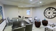This single-floor space offers a thoughtfully designed interior that seamlessly blends functionality with contemporary style. The room is finished in warm neutral tones, with smooth walls and recessed lighting that create a bright, welcoming atmosphere throughout.

A formal dining area anchors one side of the space, featuring a dark espresso-finished rectangular table paired with coordinating upholstered chairs, providing an ideal setting for meetings or communal dining. Adjacent to this, a comfortable lounge area invites relaxed conversation, centered around a pair of patterned accent chairs with coordinating throw pillows and a low coffee table. Sculptural dark wood side tables add an artistic touch and a sense of depth to the seating arrangement.

The flooring features a light-toned area rug with subtle blue detailing, tying the two zones together cohesively. A windowed pass-through or interior window brings in additional ambient light and adds architectural interest to the back wall. Tasteful decorative accents, including a ceramic vase and a glass accent piece, lend a polished, curated feel to the overall design. The space strikes a balance between elegant and approachable, making it well-suited for both social gatherings and quiet, purposeful use.