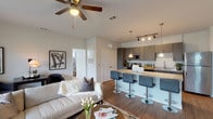 This single-floor residence showcases a thoughtfully designed open-concept layout where the living, dining, and kitchen areas flow seamlessly together. The living room is anchored by hardwood-style flooring, a ceiling fan, and large sliding glass doors that open to a private patio overlooking a lush green lawn, filling the space with abundant natural light. A dedicated workspace area sits adjacent to the living space, maintaining the open, airy feel.

The kitchen is well-appointed with slate-gray cabinetry extending to the ceiling, a full suite of stainless steel appliances including a range, microwave, dishwasher, and refrigerator, and granite countertops throughout. A spacious island with bar seating and pendant lighting above serves as a natural gathering point, complemented by track lighting overhead.

The bedrooms are carpeted for comfort, each featuring ceiling fans or wall-mounted lighting and multiple windows that allow natural light to filter in. The primary bedroom includes a generous walk-in closet outfitted with wire shelving and a window for natural light.

The bathroom features a dark-toned vanity with granite countertops, a vessel sink, and a tub-shower combination, with hardwood-style flooring that ties the space to the rest of the home.
