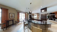 This single-floor space showcases a sophisticated and contemporary design centered around a striking curved bar with a dark granite countertop and mosaic tile base. The bar serves as the focal point of the room, complemented by sleek dark wood cabinetry, a mirrored back bar display, and polished pendant lighting that hangs at varying heights to create visual depth. Bar-height seating lines the counter, offering a social and functional gathering area.

The flooring features a terrazzo-style surface that adds texture and a refined aesthetic throughout the space. Recessed lighting is distributed across the white ceiling, ensuring consistent illumination alongside the decorative pendant fixtures.

Toward the perimeter of the space, large floor-to-ceiling windows dressed with warm rust-colored curtains allow generous natural light to filter in while offering views of an outdoor seating area. A casual seating arrangement with upholstered chairs and small tables occupies this windowed zone, providing a relaxed counterpoint to the bar area.

The overall ambiance balances modern elegance with comfort, making the space well-suited for social gatherings and hospitality-focused use.