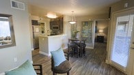 This single-story residence showcases an open-concept design that integrates the kitchen, dining, and living areas into one cohesive space. Rich, dark hardwood-style flooring runs throughout, providing a warm and grounded aesthetic that contrasts elegantly with the neutral beige walls and white trim.

The kitchen features dark cabinetry with a mosaic tile backsplash, stainless steel appliances including a refrigerator and microwave, and a peninsula with a light-colored countertop that serves as both a workspace and a casual bar-style seating area. A flush-mount ceiling light brightens the kitchen efficiently.

The dining area is positioned just off the kitchen and is anchored by a round glass-top table with dark wood chairs, illuminated by a stylish pendant light overhead. A large piece of abstract wall art adds a pop of color and visual interest to the space.

The living area flows naturally from the dining zone, furnished with comfortable seating accented by a light blue throw pillow, adding a subtle touch of color. A textured popcorn ceiling is consistent throughout, and a hallway leads to additional rooms beyond, suggesting a functional and well-organized floor plan.