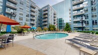 This impressive outdoor amenity courtyard is nestled within a contemporary residential complex, framed on all sides by multi-story buildings featuring a striking combination of blue-toned facades and floor-to-ceiling glass curtain walls. The architecture creates a sheltered yet open-air environment that enjoys abundant natural light throughout the day.

At the heart of the courtyard are two distinct pools: a large, oval lap pool with clearly marked swim lanes and a separate, more intimate circular pool ideal for leisure. Adjacent to the pools, a hot tub provides an additional relaxation option, surrounded by ample concrete decking lined with modern chaise lounges and colorful umbrellas for shade.

The outdoor living spaces extend well beyond the pool area. A dedicated lounge zone features wicker sectional seating, while a warm wood deck hosts a stylish outdoor seating arrangement with teal cushions centered around a fire pit — perfect for evening gatherings. Dining tables and chairs are also distributed throughout the courtyard for al fresco use.

Lush landscaping throughout includes mature evergreen trees, sculpted shrubs, seasonal flowering plants, and raised garden beds, adding vibrant greenery and natural texture that softens the urban setting. The entire courtyard is thoughtfully designed to maximize both recreation and relaxation.