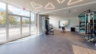 Located at 333 S Glebe Rd in Arlington, Virginia, this modern fitness center spans a single floor and is thoughtfully organized into distinct workout zones. The main exercise area features large floor-to-ceiling windows along the perimeter, flooding the space with abundant natural light and offering views of the surrounding outdoor scenery. Distinctive geometric, triangular pendant lighting fixtures are suspended from the ceiling, adding a contemporary architectural element to the bright, open environment.

The cardio zone is equipped with a row of Matrix treadmills positioned directly facing expansive windows, creating an energizing atmosphere for endurance training. Adjacent to this, the strength training area houses a variety of cable machines, weight stations, and free weights, set against warm terracotta-orange accent walls. An elliptical machine and additional resistance equipment round out the strength offerings.

A dedicated studio room, finished with hardwood-style flooring and bold orange walls, features floor-to-ceiling mirrors, a wall-mounted ballet barre, and spin bikes — well-suited for cycling classes or group fitness activities. The entrance area offers generous open floor space with carpeted and hard-surface flooring zones, providing versatility for stretching or functional training. Overall, the fitness center presents a clean, contemporary aesthetic with a well-balanced mix of cardio, strength, and studio amenities.