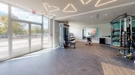 Located at 333 S Glebe Rd in Arlington, Virginia, this modern fitness center spans a single floor and is thoughtfully organized into distinct workout zones. The main exercise area features large floor-to-ceiling windows along the perimeter, flooding the space with abundant natural light and offering views of the surrounding outdoor scenery. Distinctive geometric, triangular pendant lighting fixtures are suspended from the ceiling, adding a contemporary architectural element to the bright, open environment.

The cardio zone is equipped with a row of Matrix treadmills positioned directly facing expansive windows, creating an energizing atmosphere for endurance training. Adjacent to this, the strength training area houses a variety of cable machines, weight stations, and free weights, set against warm terracotta-orange accent walls. An elliptical machine and additional resistance equipment round out the strength offerings.

A dedicated studio room, finished with hardwood-style flooring and bold orange walls, features floor-to-ceiling mirrors, a wall-mounted ballet barre, and spin bikes — well-suited for cycling classes or group fitness activities. The entrance area offers generous open floor space with carpeted and hard-surface flooring zones, providing versatility for stretching or functional training. Overall, the fitness center presents a clean, contemporary aesthetic with a well-balanced mix of cardio, strength, and studio amenities.
