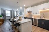 This thoughtfully designed single-floor residence combines modern aesthetics with functional living. The open-concept kitchen serves as a focal point, showcasing a striking two-tone cabinetry scheme of crisp white upper cabinets and deep navy lower cabinets, complemented by a white quartz waterfall island with bar seating. Stainless steel appliances, under-cabinet lighting, a subway tile backsplash, and pendant lights overhead complete the kitchen's polished look. Light wood-look flooring flows seamlessly from the kitchen into the living area, which benefits from multiple windows dressed in neutral linen drapes, allowing an abundance of natural light to fill the space. The living room offers a relaxed, open layout with ample room for comfortable furnishings. The bedrooms are carpeted and well-proportioned, each featuring windows with garden views and thoughtful details such as neutral wall tones and generous natural light. The bathroom continues the navy and white design language seen in the kitchen, featuring a wide vanity with dual sinks, a large mirror, and light wood-look flooring that ties the space together. Throughout the residence, the consistent use of contemporary finishes, neutral tones, and natural light creates a cohesive, inviting atmosphere.