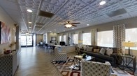 This single-floor space makes an immediate impression with its extraordinary decorative drop ceiling, adorned with a bold circular relief pattern that spans the entire room and serves as the space's defining architectural feature. A ceiling fan with a light fixture is centrally mounted, complementing the recessed lighting throughout. The floor is finished in a herringbone-patterned wood-look material, adding warmth and visual texture underfoot.

The layout is divided into two functional zones that flow seamlessly into one another. On one side, a staffed workstation and reception area features multiple computer workstations, desk seating, and framed wall art, creating a professional and organized environment for staff and visitors. Large windows along the opposite wall allow generous natural light to fill the space, dressed with patterned curtains that add a decorative touch.

The lounge area is thoughtfully furnished with plush dark sofas, patterned accent chairs, a glass coffee table, and a colorful area rug, providing a comfortable and inviting setting for guests. The overall ambiance blends professionalism with approachable warmth, making this space well-suited for a leasing office, community center, or client-facing business environment.