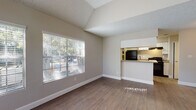 This single-story space showcases a thoughtfully designed open-concept layout that seamlessly connects the living and dining areas to the kitchen. The living area is a standout feature, boasting vaulted ceilings with a textured finish and a ceiling fan, creating an airy, spacious feel. Large windows dressed with horizontal blinds flood the space with natural light and offer views of the surrounding greenery outside.

The wood-style flooring extends throughout the main living areas, providing a clean and cohesive aesthetic. The kitchen is efficiently designed with white cabinetry, stone-look countertops, a tile backsplash, and black appliances including a refrigerator and electric range. A pass-through bar counter opens the kitchen to the living space, enhancing the sense of connectivity. In-unit laundry hookups are also visible within the kitchen area.

The bedroom is carpeted for comfort and includes a window for natural light. The bathroom features a white vanity with a stone-look countertop, a large mirror, and a tub-shower combination with white tile surround. Neutral tones throughout the space create a calm and versatile ambiance suitable for a range of furnishing styles.