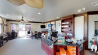 This golf pro shop offers a thoughtfully organized retail environment designed to serve golfers with convenience and comfort. The space is anchored by a dark wood-trimmed service counter with a glass display case, showcasing golf accessories and equipment available for purchase. Adjacent to the counter, built-in wooden shelving units display a curated selection of golf merchandise, including balls, apparel, and accessories, neatly organized for easy browsing.

A refrigerated beverage cooler and snack display provide refreshment options for golfers before or after their round. A wall-mounted flat-screen TV adds to the welcoming atmosphere of the space. The checkout area is fully equipped with a point-of-sale system, making transactions seamless.

Natural light filters in through a glass door entrance, brightening the carpeted interior and creating an open, airy feel. Recessed lighting throughout the ceiling ensures the entire space is well-lit. A decorative ceiling fan adds a casual, relaxed touch to the ambiance.

A small seating area with chairs and a table provides a comfortable spot for guests to relax or wait. Crown molding along the ceiling adds a polished, refined architectural detail. The overall atmosphere is clean, organized, and inviting — ideal for golfers looking to gear up or grab essentials for their game.